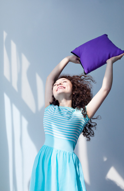 Portrait of a beautiful young girl with brown curly hair. Play with pillows. To arrange a pillow fight. Fun and joy. Purple pillow in the hands of a girl in a blue dress. - Foto, Bild