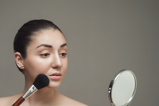 Hermosa chica mirando en el espejo y la aplicación de cosméticos con un cepillo grande. - Foto, Imagen