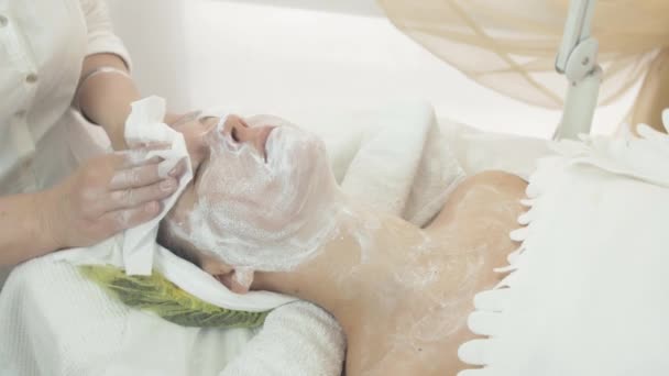 Schoonheidsspecialist veeg uit witte klei masker uit gezicht vrouw door servet in beauty salon - Video