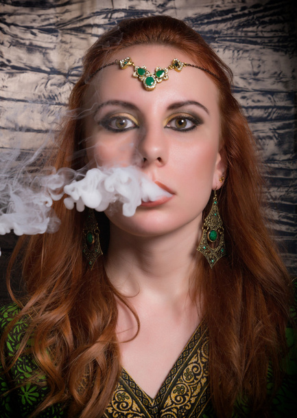 close-up portrait of a elegant young redhead woman, with oriental make-up and shawl over her head bright red lips, smokes a hookah, let the smoke out of his mouth - Foto, Imagem