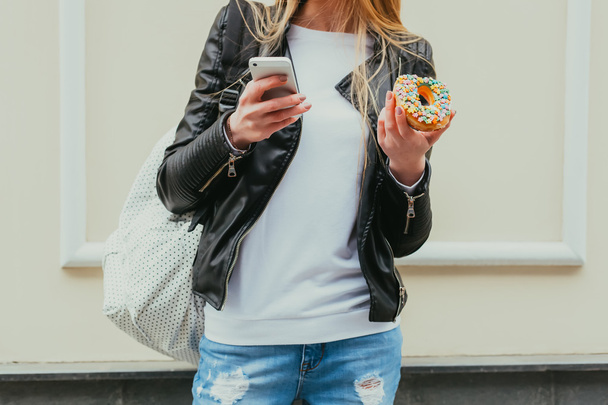 Portrait of a beautiful young sexy woman eating a donut, looks at her smart phone on the street European city. Body part. Outdoor. Close up. - Foto, afbeelding