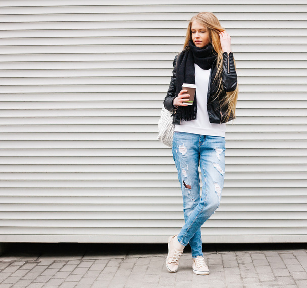 Portrait of a beautiful young blonde girl with long hair posing on a street with coffee and a backpack. Outdoor, warm color. - Valokuva, kuva