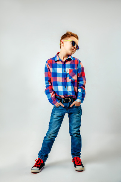 Boy wearing colorful plaid shirt, blue jeans, gumshoes, sunglasses, posing and having fun in the studio - Photo, Image