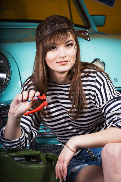 mujer atractiva joven en una gorra de béisbol se sienta en el garaje cerca del coche retro con herramientas. Chica sosteniendo alicates
 - Foto, Imagen