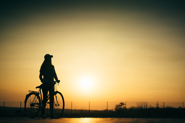 schöne Hipster-Mädchen mit einem Fahrrad auf der Straße während des Sonnenuntergangs - Foto, Bild