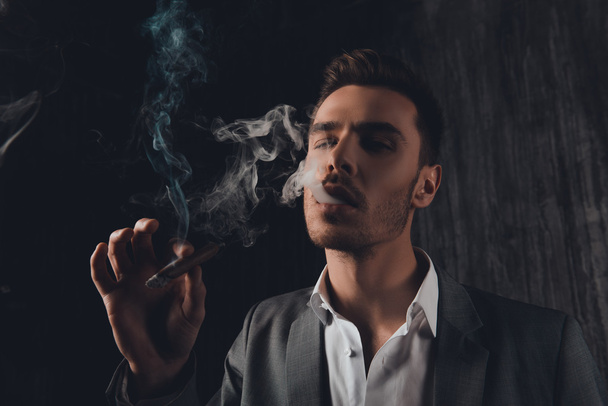 Young attractive man with cigar posing in smoke - Фото, изображение