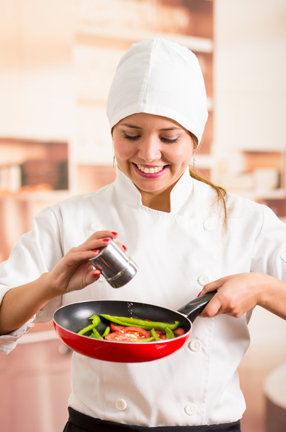 Nainen kokki tilalla punainen paistinpannu hienonnettu vihanneksia sisällä, lisäämällä suolaa metallista säiliöstä
 - Valokuva, kuva