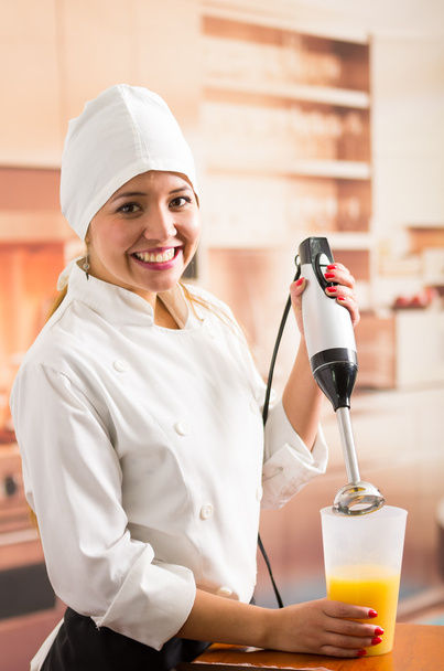 Woman chef smiling to camera using handheld electric blender with yellow liquid inside tall glass - Fotoğraf, Görsel