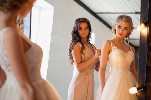 Beautiful blonde bride in luxury wedding dress and pretty bridesmaid in a morning in a loft space with a mirror and garland of lamps. Fashion modern wedding photo. - Fotó, kép