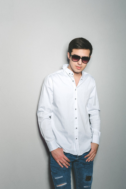 Şortlu ve beyaz gömlekli genç adam gülümsüyor. Gözlüklü duvarın yanında duruyor. - Fotoğraf, Görsel