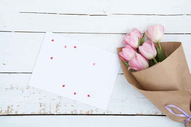 schöner Strauß rosa Tulpen neben einem weißen Umschlag und rosa Zuckerherzen auf hellem Holzhintergrund - Foto, Bild