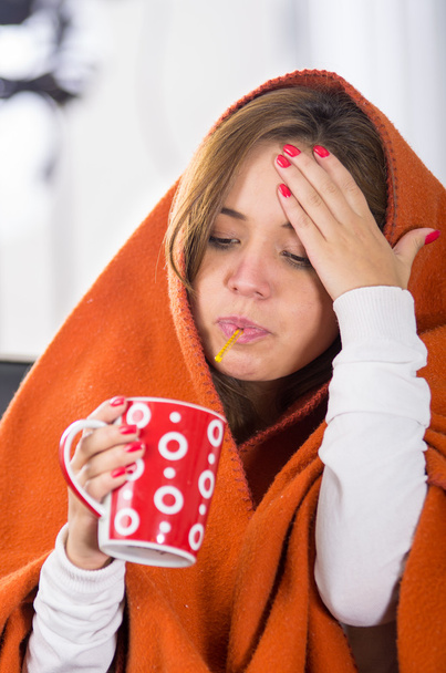 Morena sentada sob cobertor laranja, segurando copo vermelho na mão, doente com conceito de gripe
 - Foto, Imagem