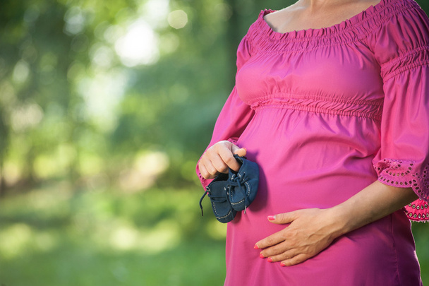 Красива вагітна жінка з дитячим взуттям стоїть на зеленому лузі
 - Фото, зображення