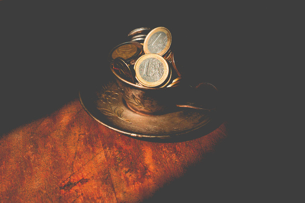 Метал, сталева чашка і блюдце з грошима євроцентом на темному тлі старого. сонячне світло
 - Фото, зображення