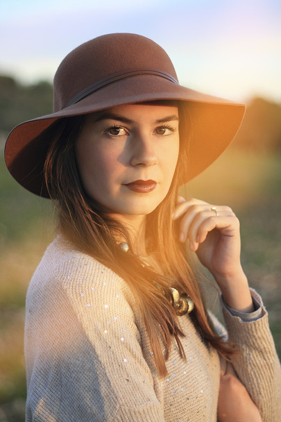 Portrait of a beautiful and distinguished young woman wearing a hat and jewelry at sunset in a open space - Φωτογραφία, εικόνα