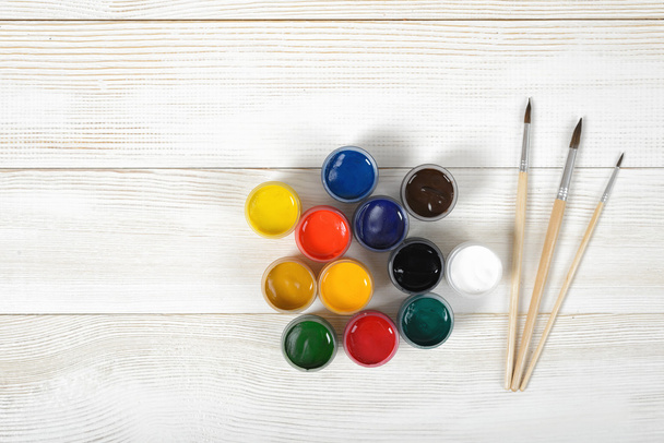 Three paint brushes and colorful gouache containers on wooden surface in top view. - Foto, imagen