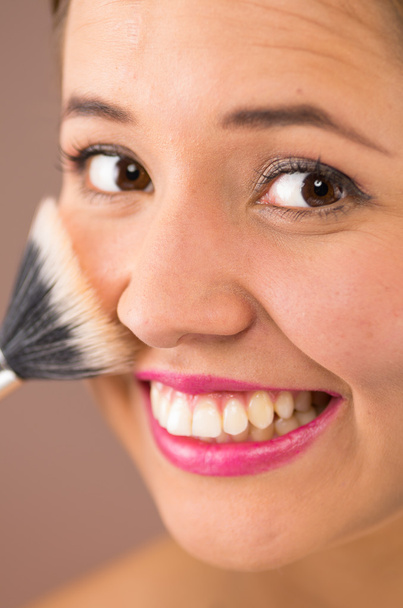 Closeup headshot young pretty hispanic woman wearing pink lipstick smiling while using dark white makeup brush on cheeks - Photo, Image