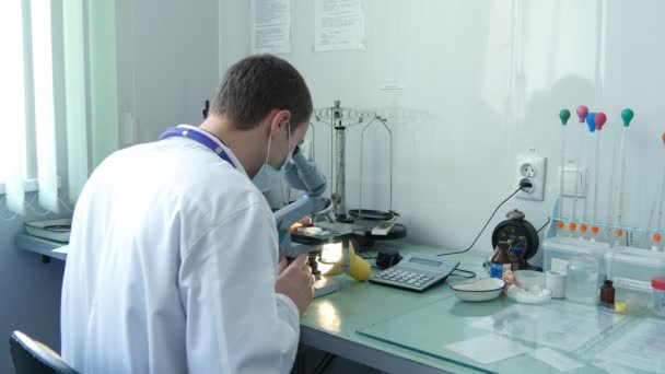 Young male doctor viewing through microscope, taking off his protective mask and smiling - Footage, Video