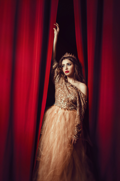 красивая стройная брюнетка женщина в желтом длинном платье пышный, с короной на голове, с одним голым плечом, стоя на фоне красного бархата занавески linny
 - Фото, изображение