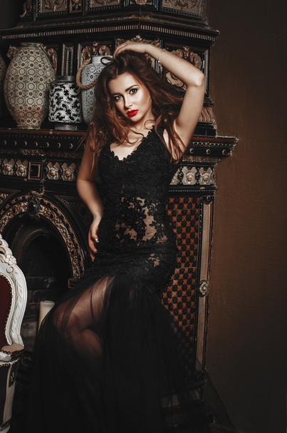 beautiful slender brunette woman in a long black lace dress stands near the ancient fireplace - Φωτογραφία, εικόνα