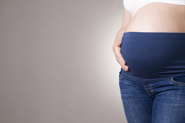 Pregnant woman in blue jeans for pregnant women on gray background. Copy space - Foto, Imagen