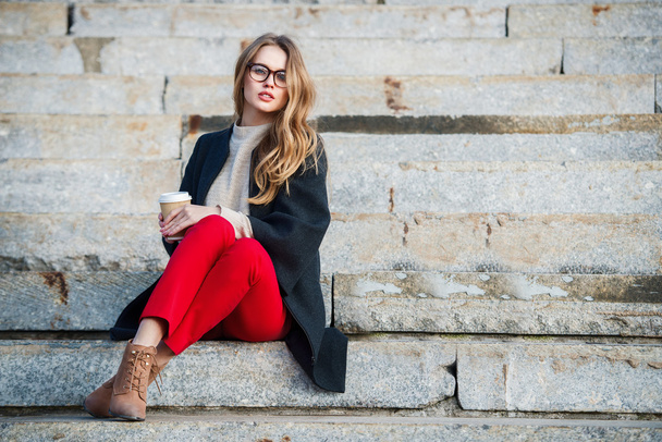 Beautiful girl with coffee cup sitting on outdoors stairs wearing red pants and glasses. - Foto, Bild