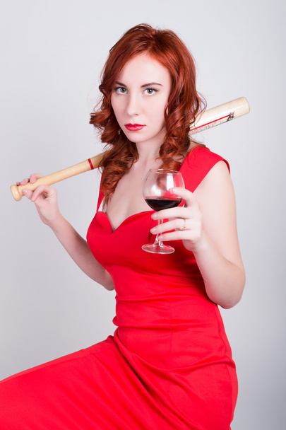 close-up portrait of Elegant young redhead woman in a red dress, drinking red wine and holding a baseball bat - Zdjęcie, obraz