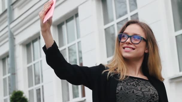 Velmi krásná a atraktivní dívka, která nosí brýle, dělá Selfie nedaleko školy. Žena s úsměvem a fotografoval sami na telefonu. Ona je šťastná - Záběry, video