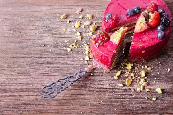 Piece of delicious raspberry cake with fresh strawberries, raspberries, blueberry, currants and pistachios on shovel, wooden background. Free space for your text. - Фото, зображення