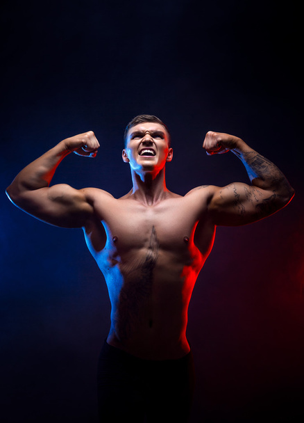 Handsome power athletic man bodybuilder. Fitness muscular body on dark smoke background. Perfect male. Awesome bodybuilder, tattoo, posing hands up. Viktory. - Photo, image