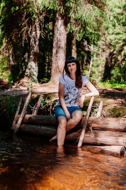 Young woman sitting on logs and lowered her feet into the river. - Foto, imagen