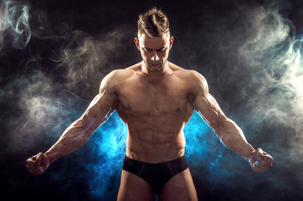 Handsome power athletic man bodybuilder. Fitness muscular body on dark smoke background. Perfect male. Awesome bodybuilder, posing. - 写真・画像