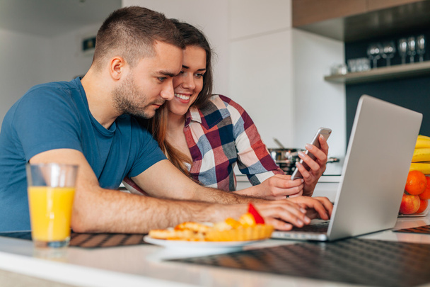 Young couple sitting in the kitchen. Man is using laptop. Girl i - Фото, изображение