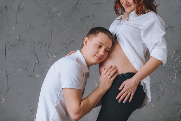 The man listens hugging the belly of pregnant women. There is a place for the text. On a gray background. She looks at him - Фото, изображение