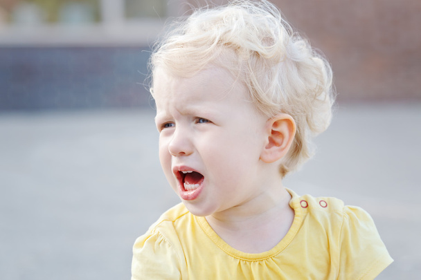 Close up portrait of crying screaming little white blond Caucasian toddler boy girl in yellow clothes outside on summer day - Photo, Image