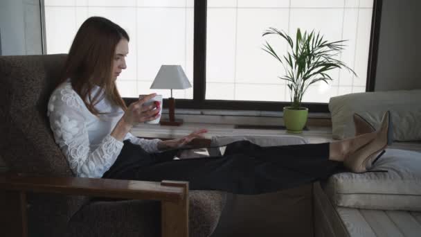 Kobieta siedzi i pije kawę jest patrząc przez jej urządzenie - Materiał filmowy, wideo