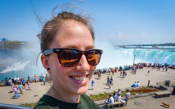 Сияющая молодая девушка счастливо улыбается, в солнечных очках, смотрит в камеру, на Ниагарский водопад в солнечный светлый день
. - Фото, изображение