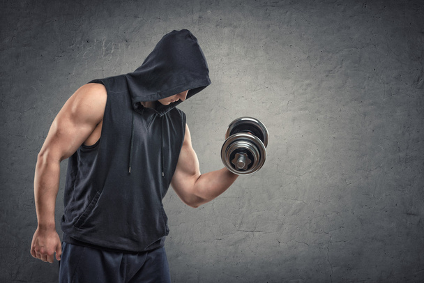 Muscular young guy in hoodie lifting a dumbbell to show his biceps - Foto, immagini