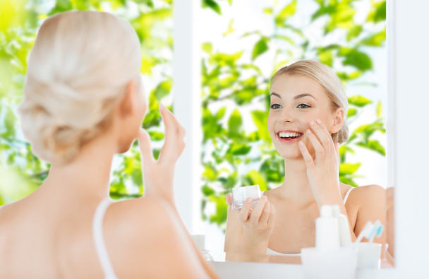 happy woman applying cream to face at bathroom - Photo, image