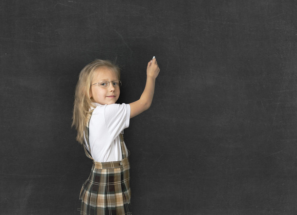 young sweet junior schoolgirl with blonde hair smiling happy writing with chalk in blackboard - Fotó, kép