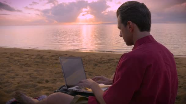 Człowiek działa na jego laptopie na plaży podczas Wschodu lub zachodu słońca - Materiał filmowy, wideo