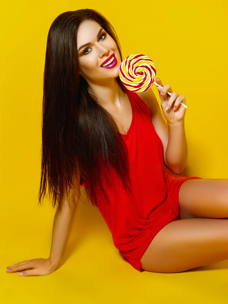 Krásná mladá dívka s candy v jeho rukou, usmíval se a pózuje pro fotografie, červené tričko, krásné a zdravé vlasy, žluté pozadí - Fotografie, Obrázek