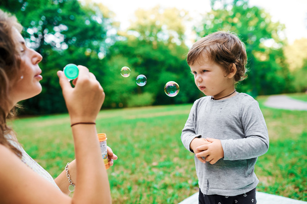 Young pregnant mother resting with her son in summer park and blow bubbles - Foto, imagen