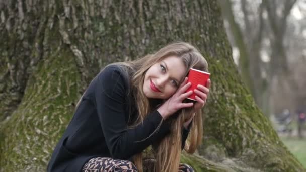 Young stylish blonde woman drinking coffee to go in the autumn park. Leafless tree on background - Video