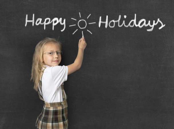сладкая младшая школьница с светлыми волосами стоя счастливый и улыбаясь писать с мелом счастливые праздники
 - Фото, изображение