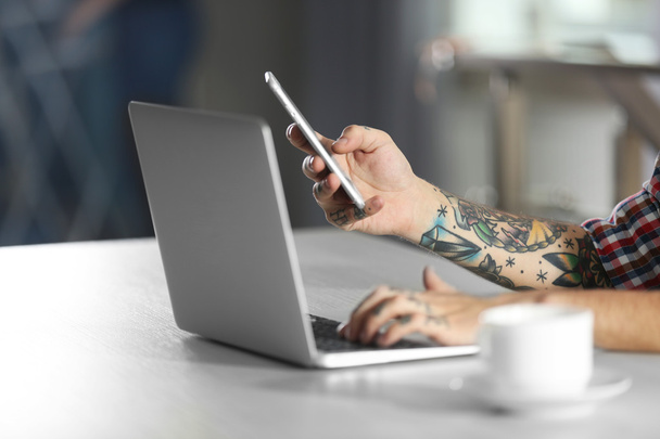 Hombre joven con tatuaje usando el ordenador portátil y el teléfono móvil en la mesa
 - Foto, Imagen