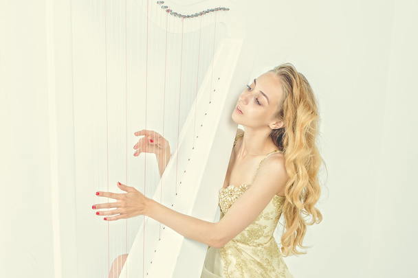白い背景に、ハープを弾く長いブロンドの髪とゴールドのドレスでエレガントな女性 - 写真・画像
