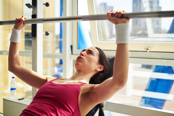 Woman is pressing a weights upwards in gym near window - Fotoğraf, Görsel
