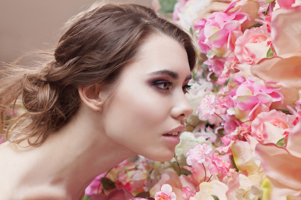 Portrait of  beautiful fashion girl, smelling a many flowers. Beautiful makeup and messy romantic hairstyle. Flowers background. Green eyes. - Φωτογραφία, εικόνα