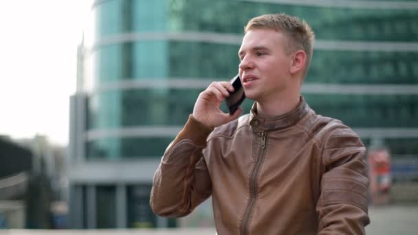 young man wearing a leather jacket talks on the phone in downtown slow motion - Materiał filmowy, wideo